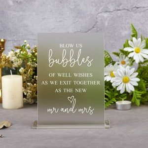 May include: A clear acrylic sign with white text that reads "Blow us bubbles of well wishes as we exit together as the new mr and mrs". The sign is sitting on a table with white flowers and candles.