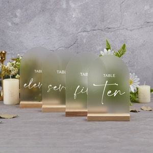 May include: Four frosted acrylic table number signs with wooden bases. The numbers are written in cursive and are eleven, seven, fifteen, and ten.