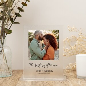 Puede incluir: Un marco de fotos de acrílico transparente con fondo esmerilado. El marco presenta una foto de una pareja abrazándose, con el texto "The best is yet to come" y "Alex + Delaney Engaged 08.05.2023" impreso debajo de la foto.