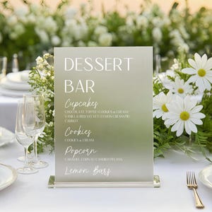 May include: A frosted acrylic dessert bar sign with the words "DESSERT BAR" in white. Below, the sign lists cupcakes, cookies, popcorn, and lemon bars. The sign is on a white table with floral arrangements.