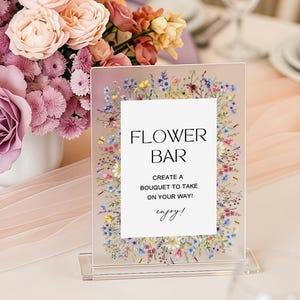 Könnte beinhalten: Ein Blumenarrangement mit violetten, rosafarbenen und pfirsichfarbenen Rosen und Rittersporn in einer weißen Vase, neben einem klaren Acrylschild mit Blumenrand, auf dem FLOWER BAR, CREATE A BOUQUET TO TAKE ON YOUR WAY, enjoy! steht.