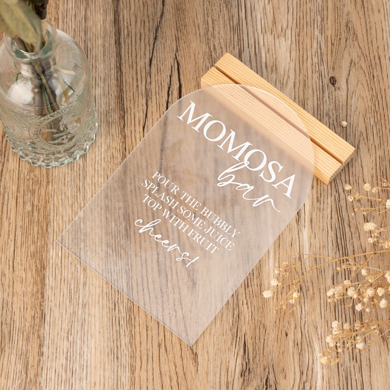 May include: A clear acrylic sign with the text "Mimosa Bar" in white cursive font. The sign also includes the instructions "Pour the bubbly, splash some juice, top with fruit, cheers!" in white sans-serif font.