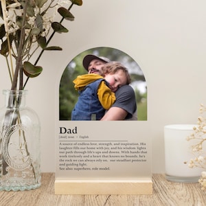 May include: A clear acrylic photo frame with a wooden base. The frame is shaped like an arch and features a photo of a father and child hugging. The frame has a definition of the word "Dad" printed on it, describing a father as a source of love, strength, and inspiration.