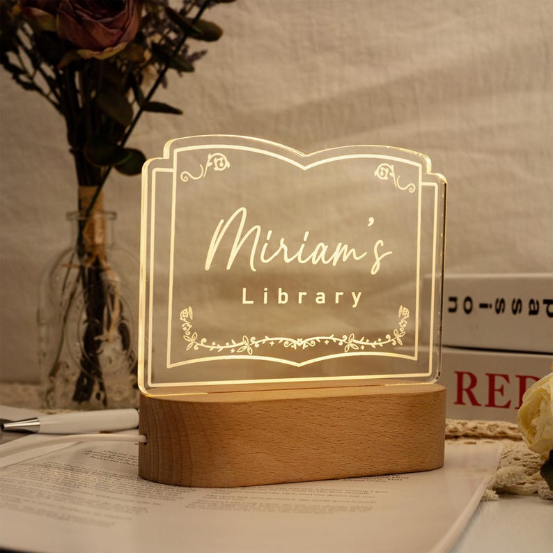 May include: A wooden base with a clear acrylic sign that reads "Miriam's Library" in gold lettering. The sign is shaped like an open book with a floral design.