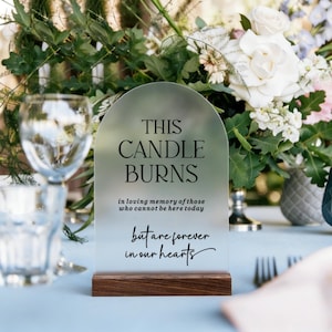 May include: An arched, frosted acrylic memorial sign with a dark wood base. The text reads: THIS CANDLE BURNS in loving memory of those who cannot be here today but are forever in our hearts. The sign is positioned upright on a light blue table.