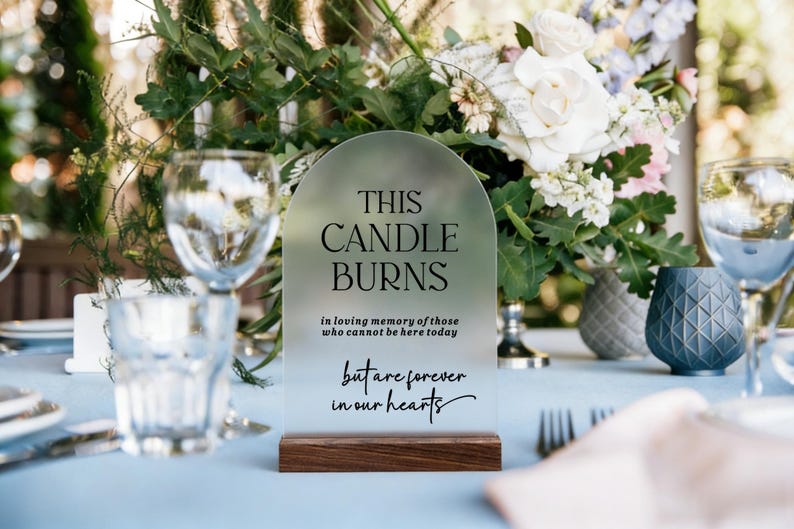 May include: An arched, frosted acrylic memorial sign with a dark wood base. The text reads: THIS CANDLE BURNS in loving memory of those who cannot be here today but are forever in our hearts. The sign is positioned upright on a light blue table.