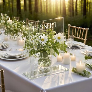 May include: A clear acrylic dessert bar sign with white text, including "Dessert Bar" and a list of treats. The sign is on a white tablecloth, with a floral arrangement and candles. The setting is outdoors.