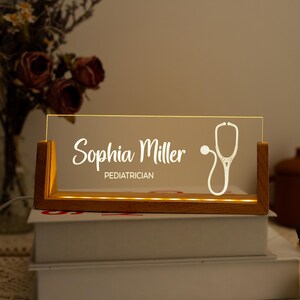Könnte beinhalten: Eine Holz-Schreibtischlampe mit einer transparenten Acrylplatte. Auf der Platte stehen der Name "Sophia Miller" und der Titel "Kinderarzt" in weißer Schrift sowie eine Stethoskop-Grafik. Die Lampe steht auf einem Stapel Bücher.