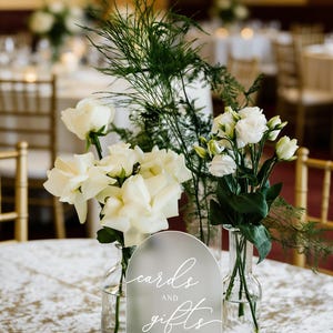 Op de afbeelding: Een bruiloft tafeldecoratie met witte rozen, groen en een mat acryl bord met de tekst "kaarten en cadeaus". Het bord staat op een houten voet. Een kleine kaars in een glazen houder staat ook op de tafel.