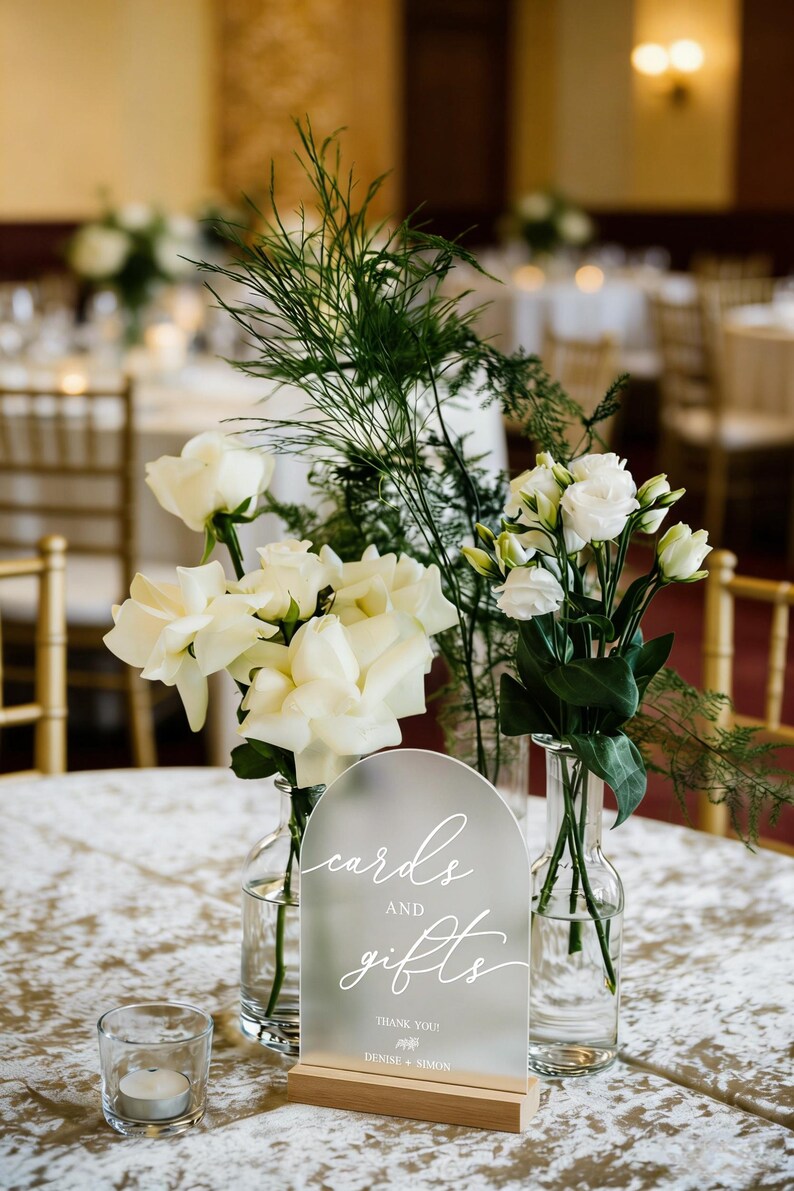 Op de afbeelding: Een bruiloft tafeldecoratie met witte rozen, groen en een mat acryl bord met de tekst "kaarten en cadeaus". Het bord staat op een houten voet. Een kleine kaars in een glazen houder staat ook op de tafel.