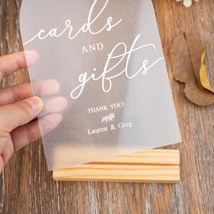 Op de afbeelding: Een helder acryl bord met de woorden "cards and gifts" in elegant wit script. Het bord bevat "Thank You!" en de namen "Lauren & Greg". Het wordt vastgehouden in een natuurlijke houten standaard.