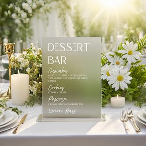 May include: A frosted glass dessert bar sign with the words "DESSERT BAR" in white. Below, the sign lists cupcakes, cookies, popcorn, and lemon bars. The sign is on a white tablecloth, with flowers and candles in the background.