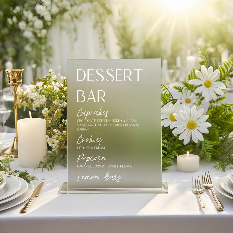 May include: A frosted glass dessert bar sign with the words "DESSERT BAR" in white. Below, the sign lists cupcakes, cookies, popcorn, and lemon bars. The sign is on a white tablecloth, with flowers and candles in the background.