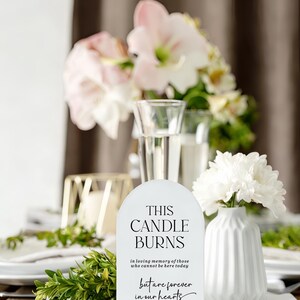May include: An arched white sign with a light wood base featuring black text that reads: THIS CANDLE BURNS in loving memory of those who cannot be here today but are forever in our hearts. The sign is positioned upright on a white surface.