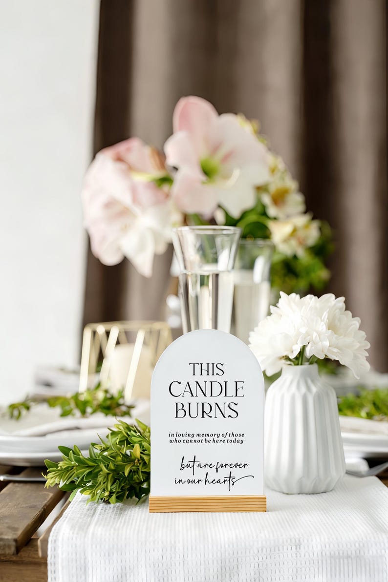 May include: An arched white sign with a light wood base featuring black text that reads: THIS CANDLE BURNS in loving memory of those who cannot be here today but are forever in our hearts. The sign is positioned upright on a white surface.