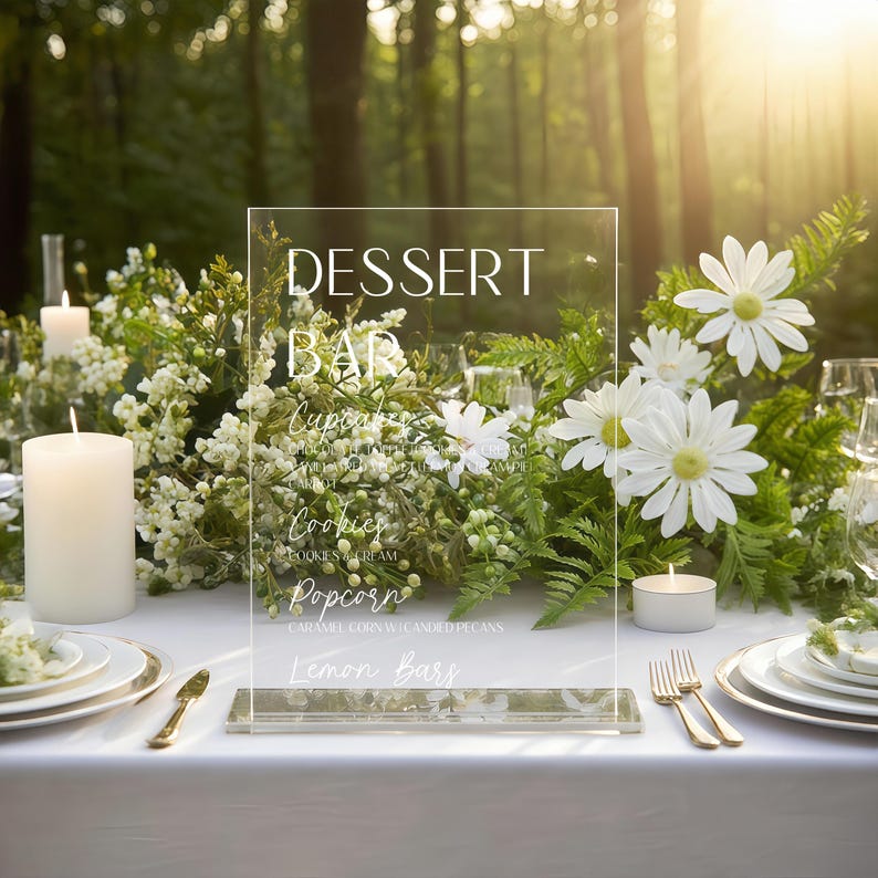 May include: A clear acrylic dessert bar sign with white text, including "Dessert Bar," and menu items like cupcakes and cookies. The sign is on a white tablecloth, surrounded by white flowers, candles, and gold-rimmed plates. The background is a blurred forest.