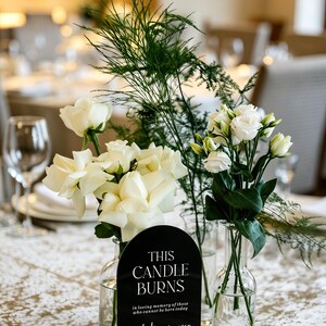 May include: A black arched acrylic sign with white text reading "THIS CANDLE BURNS in loving memory of those who cannot be here today but are forever in our hearts" set in a light wood base, flanked by two clear glass vases with white flowers.