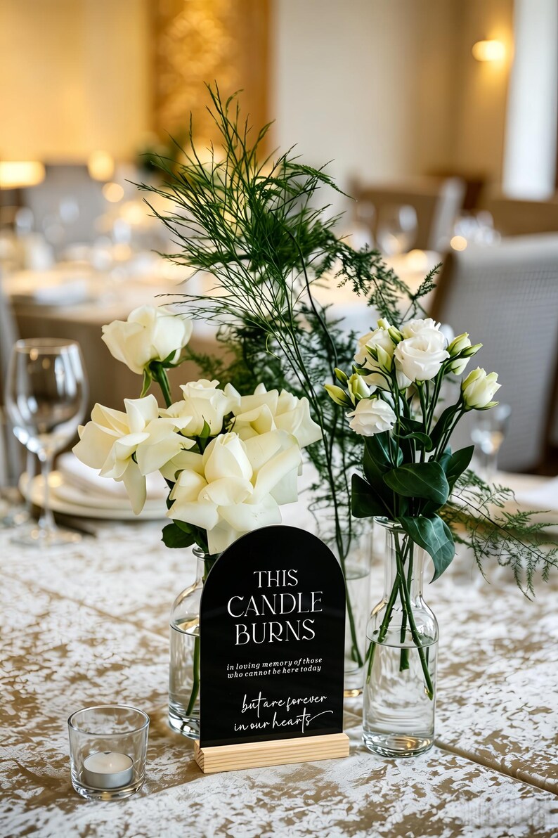 May include: A black arched acrylic sign with white text reading "THIS CANDLE BURNS in loving memory of those who cannot be here today but are forever in our hearts" set in a light wood base, flanked by two clear glass vases with white flowers.