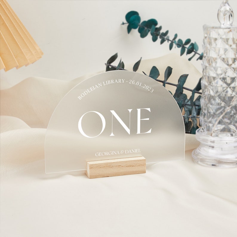 Puede incluir: Un n&uacute;mero de mesa de acr&iacute;lico esmerilado con la palabra "ONE" en texto blanco. El n&uacute;mero de mesa tiene la forma de un semic&iacute;rculo y est&aacute; sobre un soporte de madera. El texto "BODLEIAN LIBRARY - 26.01.2023" y "GEORGINA & DANIEL" tambi&eacute;n est&aacute;n impresos en el n&uacute;mero de mesa.