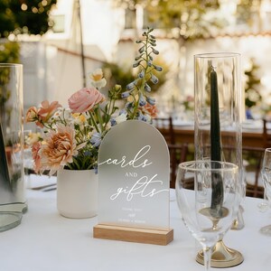 Op de afbeelding: Een helder acrylbord met de woorden "cards and gifts" in elegant wit schrift, op een houten voet. Het bord maakt deel uit van een bruiloftsreceptietafelsetting, met bloemen en glaswerk op de achtergrond.