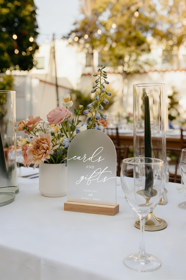 Op de afbeelding: Een helder acrylbord met de woorden "cards and gifts" in elegant wit schrift, op een houten voet. Het bord maakt deel uit van een bruiloftsreceptietafelsetting, met bloemen en glaswerk op de achtergrond.