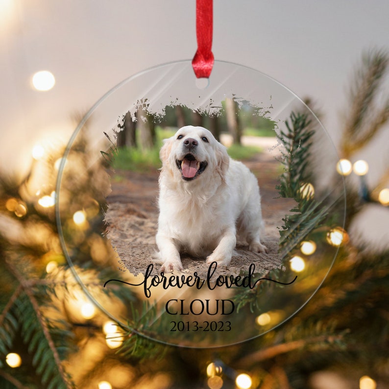 May include: A round glass ornament with a photo of a white dog. The ornament has the text "forever loved" written in cursive, followed by the name "CLOUD" and the dates "2013-2023".