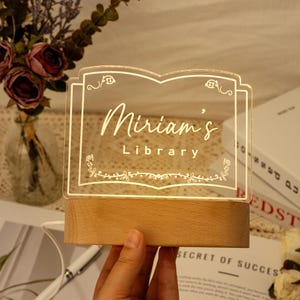 May include: A white acrylic sign shaped like a book with a wooden base. The sign reads "Miriam's Library" with a decorative floral design.
