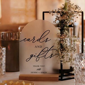 Op de afbeelding: Een mat acryl bord met de woorden "kaarten en cadeaus" in elegant schrift, geplaatst op een houten voet. Het bord bevat de namen Denise en Simon. De tafel is versierd met bloemen en kaarsen.