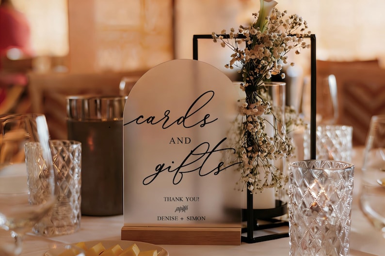 Op de afbeelding: Een mat acryl bord met de woorden "kaarten en cadeaus" in elegant schrift, geplaatst op een houten voet. Het bord bevat de namen Denise en Simon. De tafel is versierd met bloemen en kaarsen.