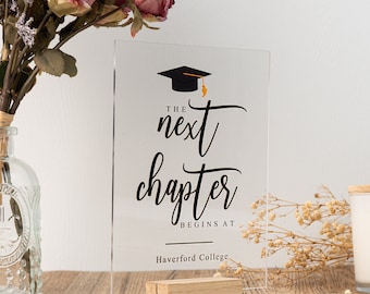 May include: A clear acrylic sign with black text that reads "The next chapter begins at Haverford College". A black graduation cap with a gold tassel is above the text. The sign is on a small wooden stand.