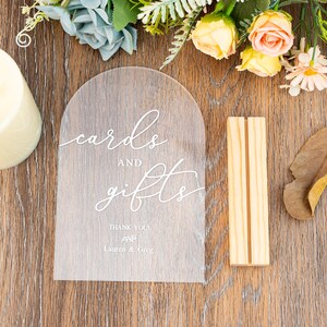 Op de afbeelding: Transparant acrylbord met de woorden "cards and gifts" in elegant wit schrift. Het bord heeft een gewelfde bovenkant en bevat de namen "Lauren & Greg" onder "Thank You!". Een houten standaard staat naast het bord, met bloemversieringen op de achtergrond.