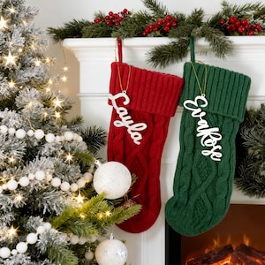 May include: Two cable-knit Christmas stockings, one red and one green, hang from a white mantelpiece. The red stocking has the name "Layla" and the green stocking has "EvaRose". A decorated Christmas tree is on the left.
