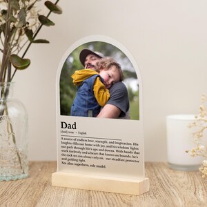 May include: A clear acrylic arch with a photo of a father and child. The arch has a white background with the word "Dad" in black text and a definition of the word. The definition reads: "A source of endless love, strength, and inspiration. His laughter fills our home with joy, and his wisdom lights our path through life's ups and downs. With hands that work tirelessly and a heart that knows no bounds, he's the rock we can always rely on, our steadfast protector and guiding light. See also: superhero, role model."