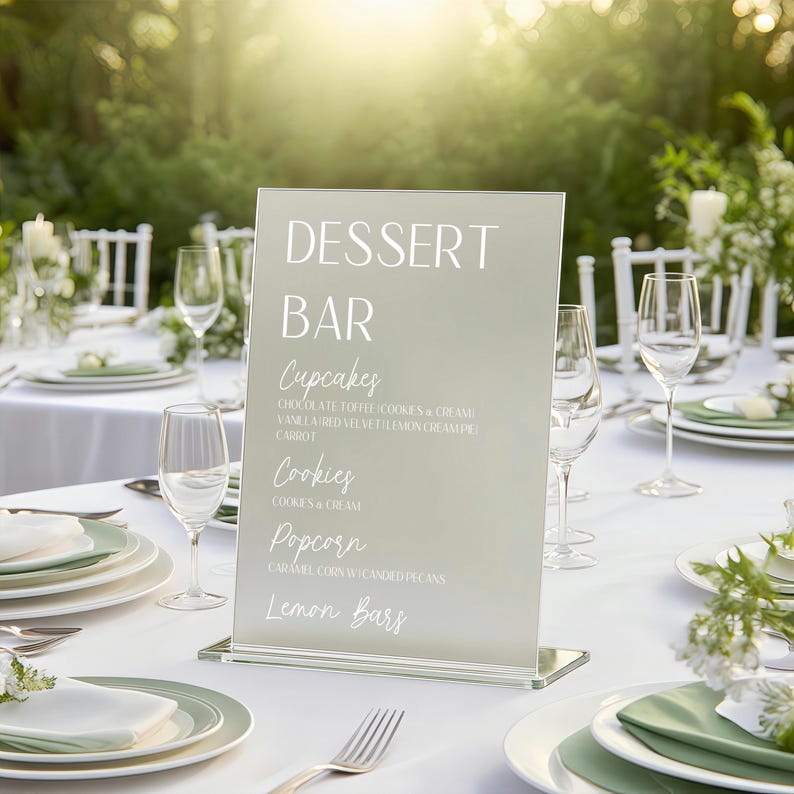 May include: A clear acrylic dessert bar sign with white text listing dessert options. The sign is displayed on a white tablecloth with plates, glasses, and silverware. The sign lists cupcakes, cookies, popcorn, and lemon bars.