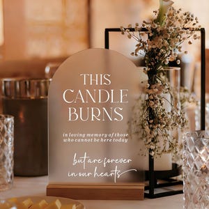 May include: An arched, frosted acrylic sign with a wooden base featuring the white text: THIS CANDLE BURNS in loving memory of those who cannot be here today but are forever in our hearts. The sign is positioned next to a black metal frame.