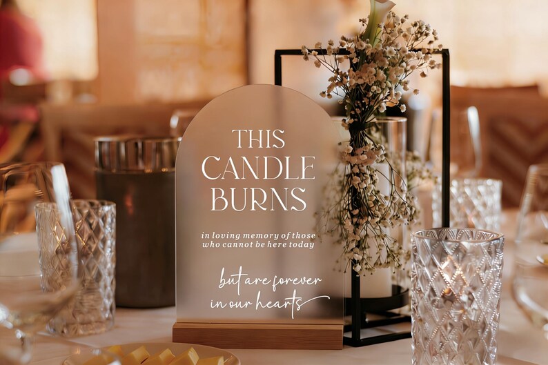 May include: An arched, frosted acrylic sign with a wooden base featuring the white text: THIS CANDLE BURNS in loving memory of those who cannot be here today but are forever in our hearts. The sign is positioned next to a black metal frame.