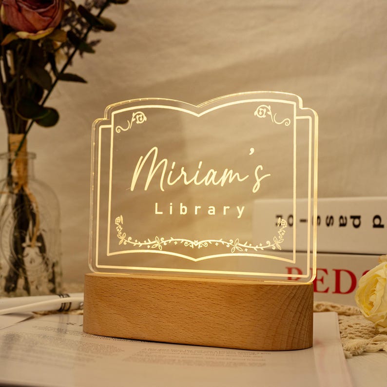 May include: A clear acrylic sign with a wooden base, shaped like an open book. The sign is illuminated from behind and features the text "Miriam's Library" in a decorative font.