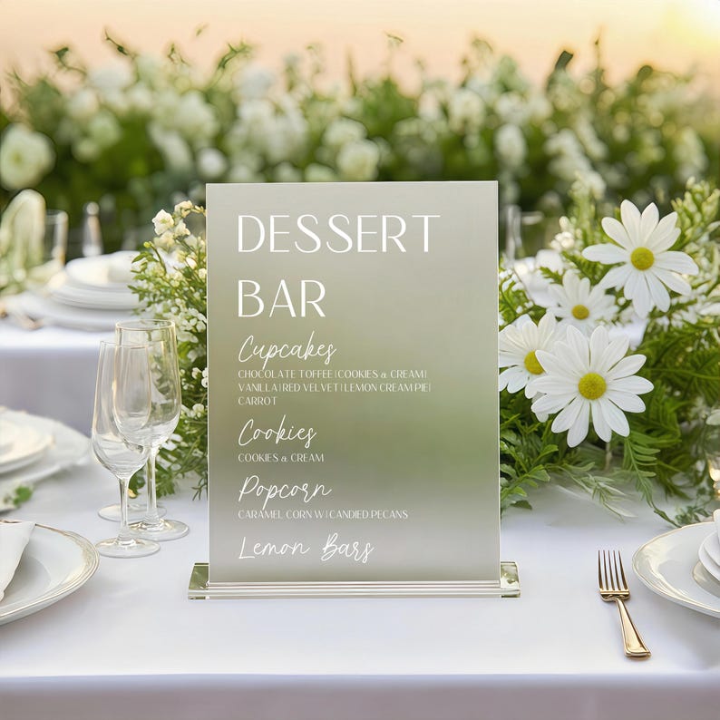 May include: A frosted acrylic dessert bar sign with the words "DESSERT BAR" in white. Below, the sign lists cupcakes, cookies, popcorn, and lemon bars. The sign is on a white table with floral arrangements.