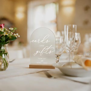 Op de afbeelding: Een helder acrylbord met de woorden "cards and gifts" in elegant wit schrift, op een houten voet. Het bord staat op een tafel met een vaas bloemen en glaswerk, geschikt voor een bruiloft of evenement.