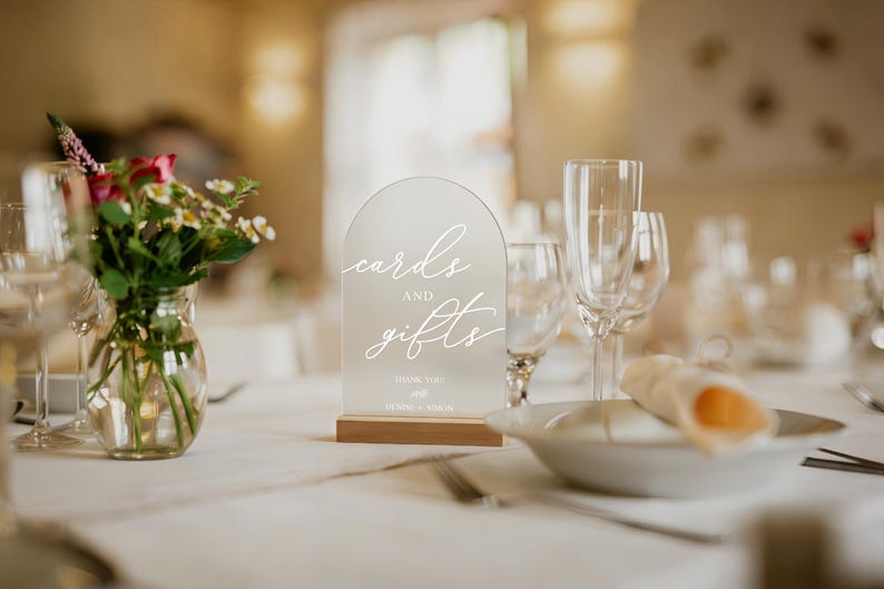 Op de afbeelding: Een helder acrylbord met de woorden "cards and gifts" in elegant wit schrift, op een houten voet. Het bord staat op een tafel met een vaas bloemen en glaswerk, geschikt voor een bruiloft of evenement.