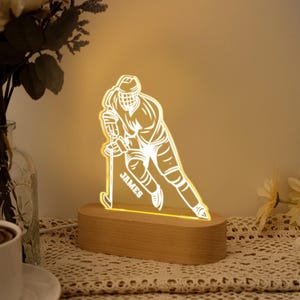 Puede incluir: Lámpara LED con forma de jugador de hockey con el nombre "JAMES" en la parte delantera. La lámpara es de acrílico transparente y se apoya en una base de madera. La lámpara emite un brillo amarillo cálido, ideal para un dormitorio o escritorio.