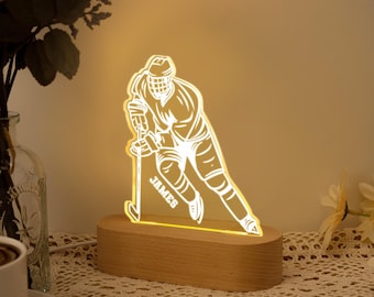 Könnte beinhalten: LED-Lampe in Form eines Hockeyspielers mit dem Namen "JAMES" auf der Vorderseite. Die Lampe besteht aus klarem Acryl und steht auf einem Holzsockel. Die Lampe strahlt ein warmes, gelbes Licht aus, ideal für ein Schlafzimmer oder einen Schreibtisch.