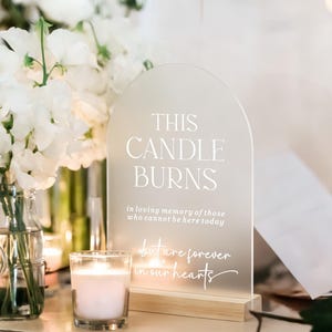 May include: An arched, frosted acrylic sign with a wooden base featuring white text that reads: THIS CANDLE BURNS in loving memory of those who cannot be here today but are forever in our hearts. The sign is displayed next to white flowers.