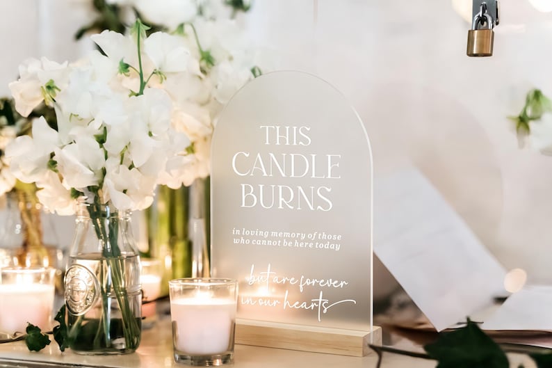May include: An arched, frosted acrylic sign with a wooden base featuring white text that reads: THIS CANDLE BURNS in loving memory of those who cannot be here today but are forever in our hearts. The sign is displayed next to white flowers.