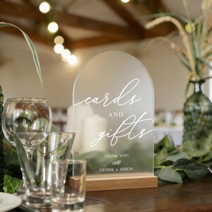 Op de afbeelding: Een helder acrylbord met de woorden "cards and gifts" in elegant wit schrift, geplaatst op een houten voet. Het bord maakt deel uit van een bruiloftstafelsetting, met groene glazen vazen en glaswerk.