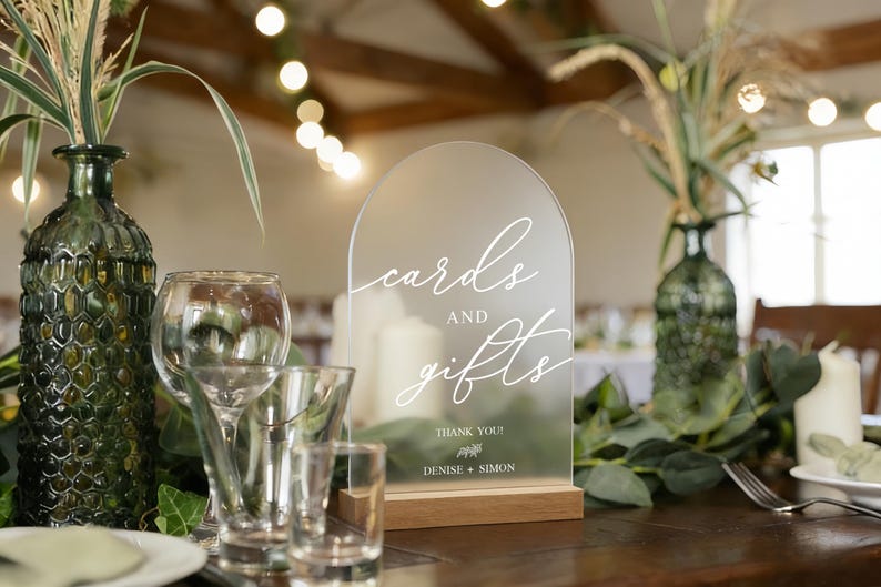 Op de afbeelding: Een helder acrylbord met de woorden "cards and gifts" in elegant wit schrift, geplaatst op een houten voet. Het bord maakt deel uit van een bruiloftstafelsetting, met groene glazen vazen en glaswerk.