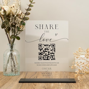 Könnte beinhalten: Ein klares Acrylschild mit schwarzem Text, der "SHARE THE love" mit einem kleinen Herzsymbol lautet. Unter dem Text befindet sich ein schwarz-weißer QR-Code mit den Anweisungen "SHARE YOUR PHOTOS WITH US! SCAN CODE AND UPLOAD!" Unter den Anweisungen steht der Text "OSCAR und NICOLE 07.18.2025".