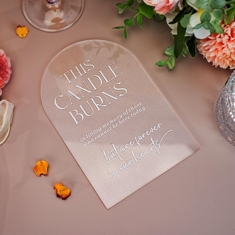 May include: An arched, semi-transparent acrylic memorial sign with white text that reads: THIS CANDLE BURNS in living memory of those who cannot be here today but are forever in our hearts. The sign has a smooth, matte finish and rounded top.
