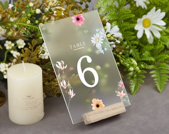 May include: A clear acrylic table number card with a floral design. The card has a wooden base and the number "6" printed in white. The text "Table" is printed in black at the top of the card.