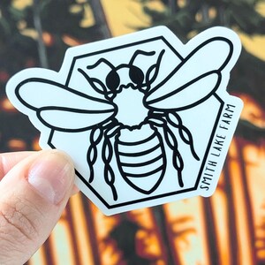 May include: A white sticker with a black outline of a bee inside a hexagon. The bee has black stripes and a white body. The text "Smith Lake Farm" is on the sticker.